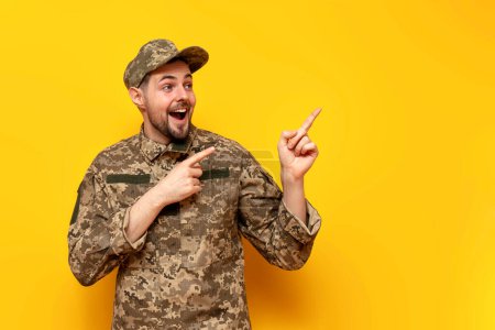 shocked young Ukrainian army soldier in camouflage pixel uniform points with hands to the side on yellow isolated background, surprised Ukrainian military cadet showing and advertising copy space in