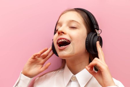 petite adolescente joyeuse avec bretelles écoute de la musique sur écouteurs et danse sur un fond rose isolé, l'enfant chante et annonce avec la bouche ouverte