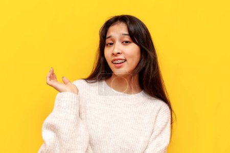young insecure asian girl with braces shrugs her shoulders over yellow isolated background, puzzled korean girl shows indifference and raises her hand