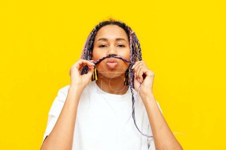 young african american woman with dreadlocks shows a mustache from her hair on a yellow isolated background, a girl with a unique hairstyle and colored braids makes faces and jokes
