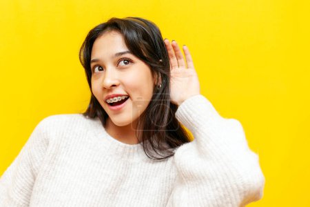 young curious asian woman with braces eavesdropping and listening on yellow isolated background, korean girl holding hand near ear, spying and wondering