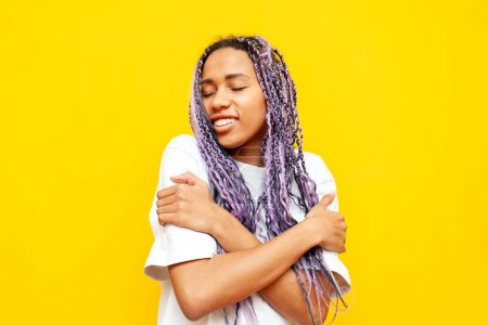 junge afrikanisch-amerikanische Frau mit Dreadlocks umarmt sich vor gelbem Hintergrund, Mädchen mit einzigartiger Frisur und farbigen Zöpfen liebt sich selbst