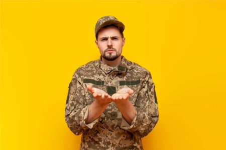 young Ukrainian army soldier in camouflage pixel uniform poking and holding empty hands on yellow isolated background, sad Ukrainian military cadet asking and showing
