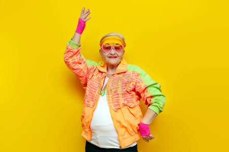verrückt lustige alte Oma in bunten Sportklamotten und rosa Brille, die auf gelbem Hintergrund tanzt, ältere Frau in jugendlichen Hipsterklamotten, die sich mit erhobenen Händen bewegt