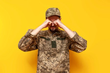unhappy offended young Ukrainian army soldier in camouflage pixel uniform crying on yellow isolated background, sad Ukrainian military cadet in depression
