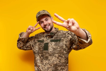 young Ukrainian army soldier in camouflage pixel uniform shows peace gesture on yellow isolated background, Ukrainian military cadet smiles and greets