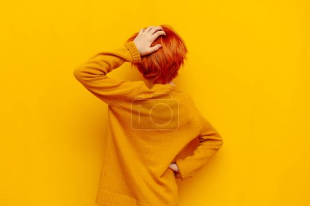 puzzled red-haired teenage boy thinks and chooses on a yellow isolated background, confused child with an orange hairstyle remembers and scratches the back of his head, rear view