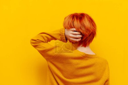 puzzled red-haired teenage boy thinks and chooses on a yellow isolated background, confused child with an orange hairstyle remembers and scratches the back of his head, rear view