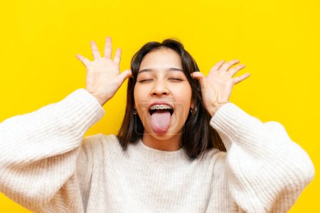 young crazy asian woman with braces shows her tongue and makes faces on a yellow isolated background, korean girl makes grimaces and jokes