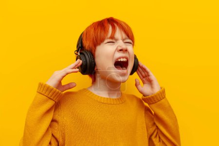 red-haired teenage boy listens to music on headphones and shouts loudly on a yellow isolated background, a child with an orange hairstyle in black headphones sings with his mouth open and announces