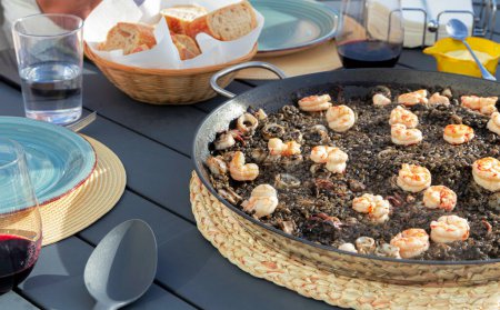 Photo for Black rice with squid and shrimps in a paella pan. Ready to eat, with bread and red wine - Royalty Free Image