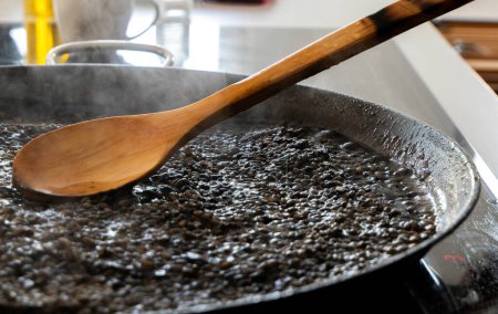 Photo for Black rice with squid being cooked in a paella pan and a wooden spoon. Black rice with cuttlefish. Rice al nero di sepia. Black risotto - Royalty Free Image