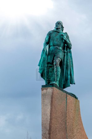 Photo for A statue of a Norse explorer with an axe is standing on a stone pedestal. The statue is green and the sky is cloudy - Royalty Free Image