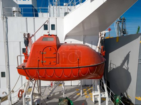 Foto de Bote salvavidas naranja en un buque de carga, para evacuación de emergencia. El barco se ve de cerca, con partes de la nave y el cielo visibles en el fondo. - Imagen libre de derechos