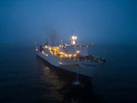 Aerial view of a ferryboat navigating through foggy conditions at night on the sea. The combination of the dark setting, the foggy conditions, and the presence of the ferryboat can create an eerie and