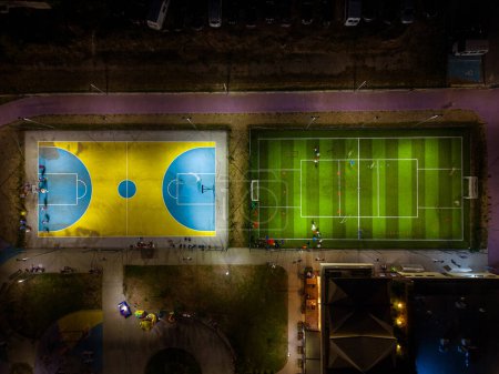 Foto de Vista aérea de un campo de deportes nocturno con campos de fútbol y baloncesto, donde los niños practican fútbol y baloncesto. - Imagen libre de derechos