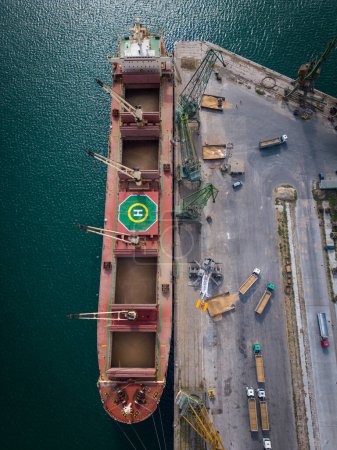 Photo for Aerial top view of a bustling seaport, where a massive cargo ship, a bulk carrier, is being loaded with wheat grains - Royalty Free Image