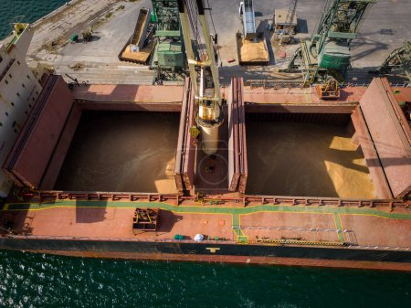 Foto de Vista aérea de primer plano de un bullicioso puerto marítimo, donde un enorme buque de carga, un granelero, está siendo cargado con granos de trigo - Imagen libre de derechos