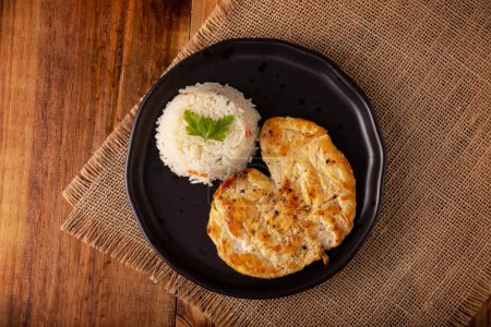 Photo for Homemade grilled chicken breast, flattened and seasoned. Accompanied by white rice served in a dark plate on a rustic wooden table. Table top view. - Royalty Free Image