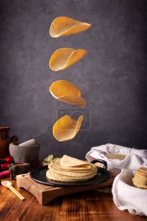 Tortillas de maíz cayendo en un comal mexicano en un entorno de cocina típica mexicana con una mesa de madera rústica y molcajetes de piedra.