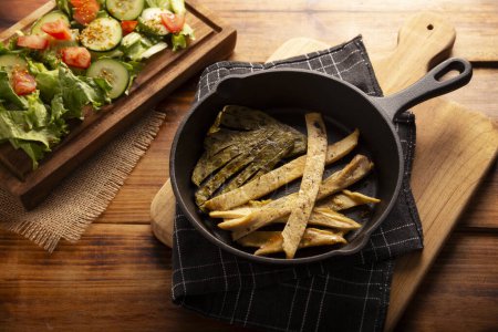 Photo for Roasted chicken breast cut into strips, roasted in a cast iron skillet accompanied by roasted nopales and fresh salad, served on a rustic wooden table. Simple and nutritious homemade recipe. - Royalty Free Image