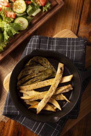 Roasted chicken breast cut into strips, roasted in a cast iron skillet accompanied by roasted nopales and fresh salad, served on a rustic wooden table. Simple and nutritious homemade recipe.