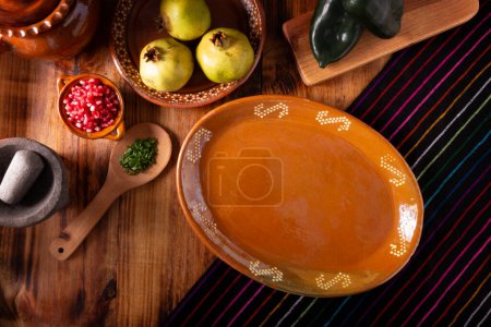 Fondo de comida mexicana. Placa tradicional de arcilla vacía sobre una mesa rústica de madera con ingredientes básicos para preparar chiles en nogada, un plato típico mexicano.