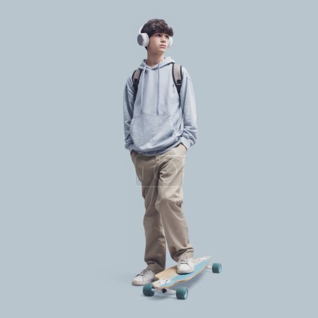 Téléchargez les photos : Adolescent cool posant avec une planche à roulettes, isolé sur fond gris - en image libre de droit