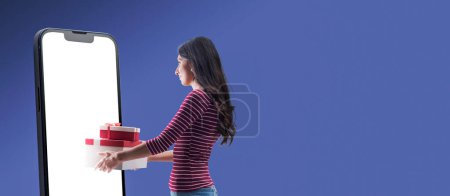 Photo for Happy woman doing online shopping on a big smartphone and receiving gifts, blank screen on the smartphone, copy space - Royalty Free Image
