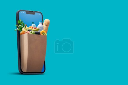 Téléchargez les photos : Magasins d'alimentation en ligne et livraison à domicile : sac rempli d'épicerie sortant d'un écran de smartphone - en image libre de droit
