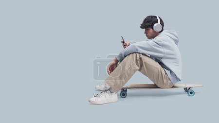 Téléchargez les photos : Skater portant des écouteurs et assis sur son patin, il se connecte à son smartphone et écoute de la musique, espace de copie vierge - en image libre de droit