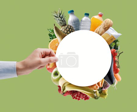 Photo for Hand holding a circular sign framed by fresh groceries, grocery shopping and nutrition concept - Royalty Free Image