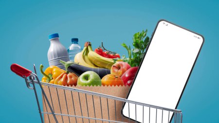 Téléchargez les photos : Supermarché panier plein d'épicerie et smartphone avec écran blanc, concept d'épicerie en ligne - en image libre de droit
