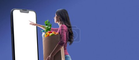 Foto de Woman holding a grocery bag and doing online grocery shopping using a big smartphone with blank screen - Imagen libre de derechos