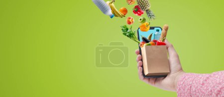 Téléchargez les photos : Woman holding her smartphone and doing online grocery shopping, she is receiving fresh food and drinks - en image libre de droit