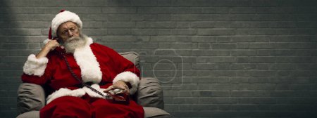Foto de Santa Claus sentado en el sillón y teniendo una llamada telefónica, está sosteniendo un receptor y escuchando - Imagen libre de derechos