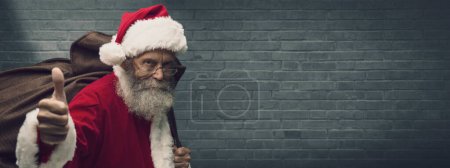 Photo for Confident happy Santa Claus giving a thumbs up and carrying Christmas gifts in a huge sack - Royalty Free Image