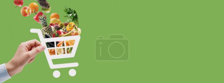 Téléchargez les photos : Main tenant une icône de chariot pleine d'épicerie fraîche : application d'épicerie et de livraison en ligne - en image libre de droit