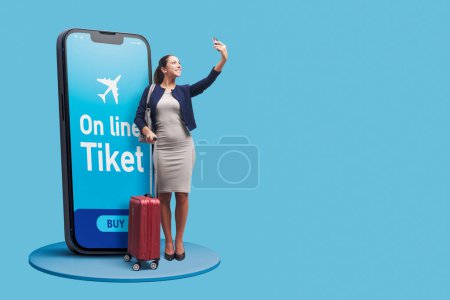 Téléchargez les photos : Femme d'affaires à la mode avec sac trolley réservant ses billets d'avion en ligne sur smartphone, elle prend un selfie - en image libre de droit