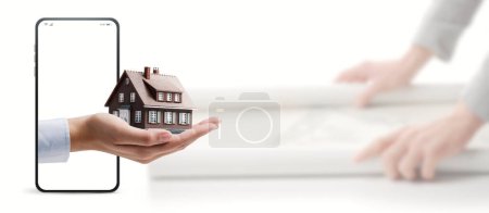 Téléchargez les photos : Architecte vérifiant un projet de maison et tenant à la main une maison modèle sur un écran de smartphone : application de rénovation et d'immobilier - en image libre de droit