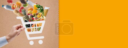 Photo for Hand holding a shopping cart icon full of fresh groceries: online grocery shopping and delivery app - Royalty Free Image