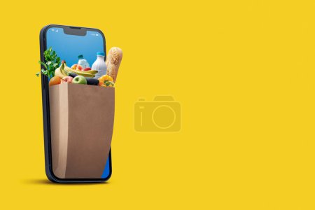 Photo for Online grocery shopping and home delivery: bag full of groceries coming out of a smartphone screen - Royalty Free Image