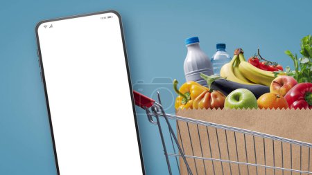 Téléchargez les photos : Supermarché panier plein d'épicerie et smartphone avec écran blanc, concept d'épicerie en ligne - en image libre de droit