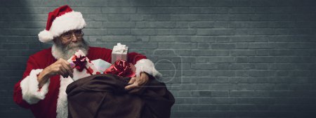 Photo for Santa Claus is bringing Christmas gifts in a huge sack, he is holding a present with a red ribbon - Royalty Free Image
