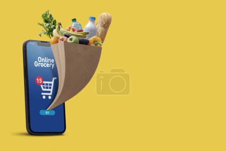 Foto de Aplicación de comestibles en línea en el teléfono inteligente y la bolsa de supermercado completa que sale de la pantalla del teléfono inteligente, espacio para copiar - Imagen libre de derechos