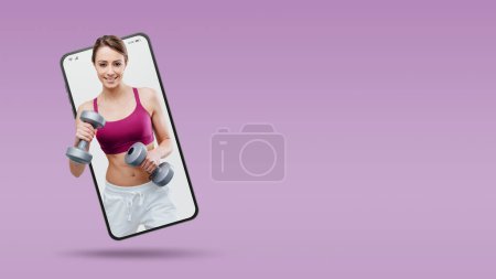 Téléchargez les photos : Smiling young sportswoman working out with dumbbells in a smartphone videocall and smiling, online  service concept - en image libre de droit