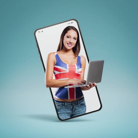 Téléchargez les photos : Smiling young girl with laptop wearing a British flag  in a smartphone videocall and smiling, online  education concept - en image libre de droit