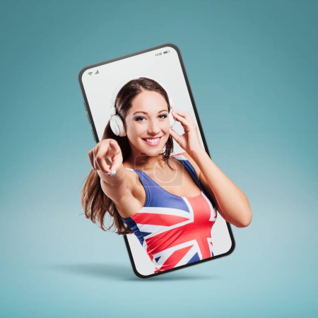 Téléchargez les photos : Cheerful pop girl wearing a British flag top, listening to music  in a smartphone videocall and smiling, online  concept - en image libre de droit