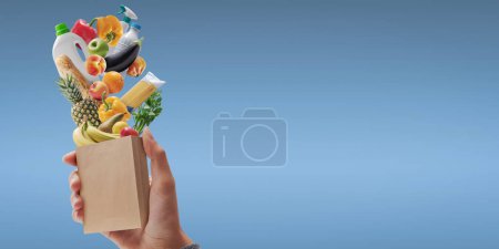 Téléchargez les photos : Customer holding a miniature paper bag with fresh groceries falling inside, grocery shopping concept - en image libre de droit