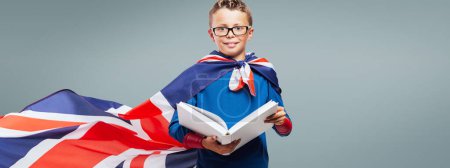 Téléchargez les photos : Superhéros mignon garçon lisant un livre, il porte un drapeau britannique comme un concept de cape, d'éducation et de plaisir - en image libre de droit
